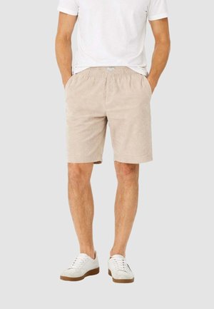 Hombre con pantalones cortos de pana beige, camiseta blanca y zapatillas blancas, de pie con las manos en los bolsillos sobre un fondo gris claro.