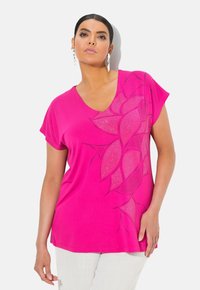 Růžové tričko s V-výstřihem a krátkými rukávy, s květinovým vzorem z malých kamínků na jedné straně. Měkká tkanina s volným střihem.