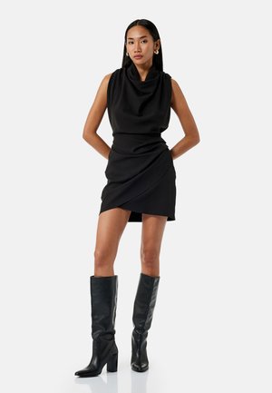 Robe noire sans manches avec un décolleté drapé et un design tombant, associée à des bottes en cuir noires hautes jusqu'au genou avec un talon carré.
