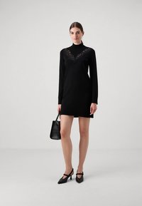 Fekete hosszú ujjú ruha csipkedíszítéssel a nyakkivágásnál, texturált fekete kézitáskával és hegyes orrú magassarkú cipővel párosítva.