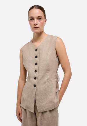 Femme portant un gilet en lin beige sans manches avec des boutons noirs et un pantalon assorti, debout devant un fond clair uni.