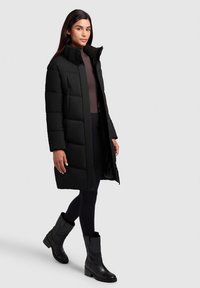 Schwarze gepolsterte Jacke mit hohem Kragen, langen Ärmeln und Reißverschluss, kombiniert mit schwarzen Ankle Boots und figurbetonten schwarzen Leggings.