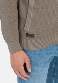 Lichtbruine gebreide trui met geribde manchetten en zoom, met een tan accent en een klein label op de linkerbenedenkant. Draagt met blauwe jeans.