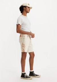 Camiseta gris claro desgastada y pantalones cortos de mezclilla beige con patrones decorativos en naranja. Zapatillas negras con suelas blancas. Diseño sencillo.