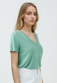 T-shirt a scollo a V di colore verde chiaro, realizzata in tessuto morbido. Maniche corte con vestibilità rilassata. Indossato con pantaloni bianchi. Design minimalista.
