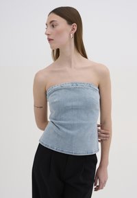 Top senza spalline in denim azzurro chiaro con un corpetto aderente e orlo grezzo. Il modello indossa pantaloni neri, con una texture liscia e un design informale.