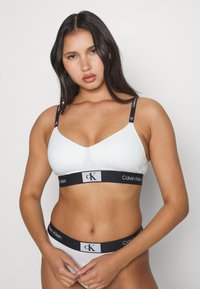 Bijela Calvin Klein braletica s tankim crnim trakama, s logotipom na dnu. Glatka, rastezljiva tkanina s jednostavnim dizajnom.