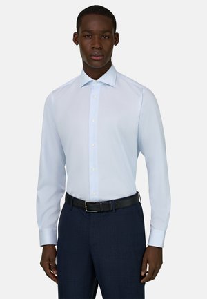 Mężczyzna ubrany w jasnoniebieską, długorękawową koszulę włożoną w ciemnoniebieskie spodnie z czarnym paskiem, stojący na jednolitym tle.