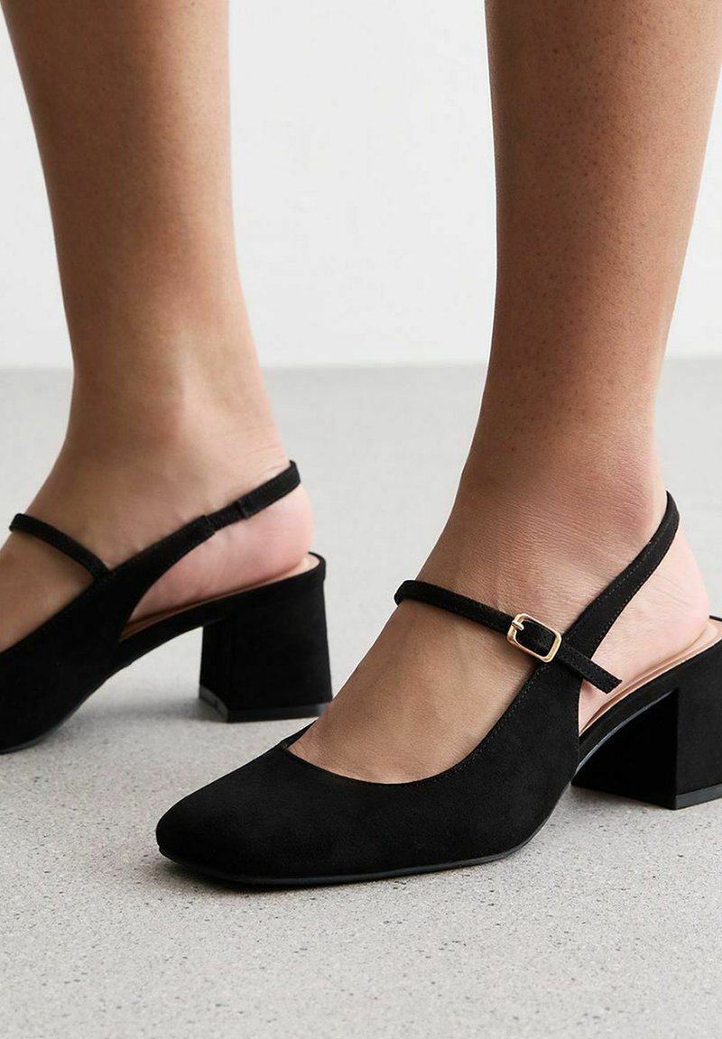 Zapatos de ante negro tipo slingback con punta afilada, correa ajustable y tacón grueso, con un acento de hebilla dorada.