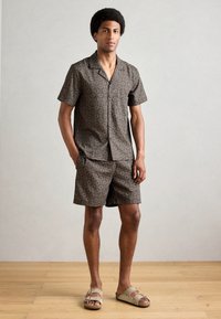 Rövid ujjú ing és sort szett barna színben, geometrikus mintával, könnyű anyagból készült. Nyitott orrú szandálokkal viselve.