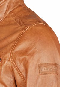Braune Lederjacke mit strukturierten Oberflächen, gesteppten Details und einem Markenpatch am linken Ärmel.