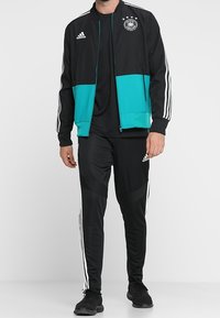 Chaqueta de chándal negra y turquesa de Adidas con rayas blancas, que cuenta con cierre de cremallera y el escudo de Alemania. Combinada con pantalones negros a juego.