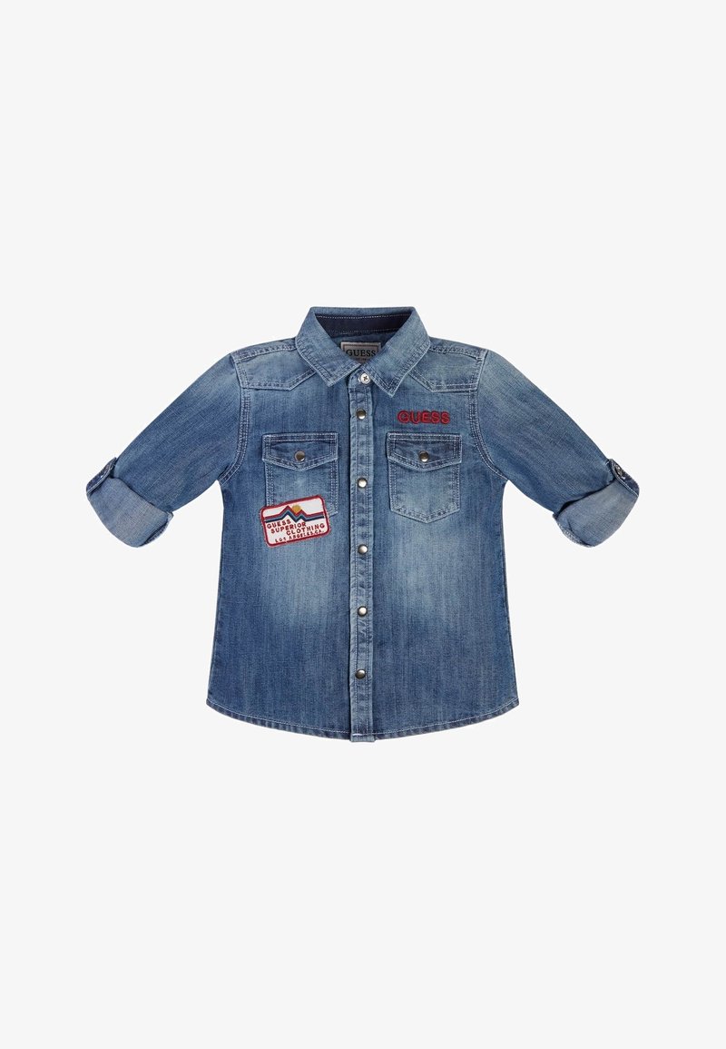 Chemise en jean bleu à boutons, manches retroussées, deux poches poitrine, un logo rouge "GUESS" et un patch coloré sur la poche gauche.