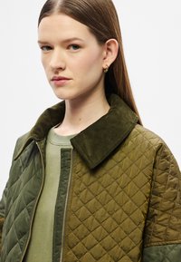 Πλισέ μπουφάν τύπου bomber σε ελαιόχρωμο πράσινο με γιακά από βελούδο κοτλέ, διαθέτει φερμουάρ στο μπροστινό μέρος και συνδυάζεται με ένα ελαφρύ πράσινο τοπ από κάτω.