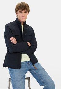 Granatowa pikowana kurtka z wysokim kołnierzem, zamkiem błyskawicznym z przodu i dwiema kieszeniami na piersi. Nosi na sobie jasną zieloną koszulę, zestawioną z jasnymi dżinsami.