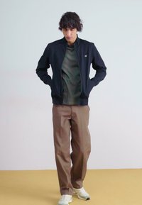 Svart glidelåsjakke over en olivengrønn skjorte, paret med brune bukser og hvite sneakers. Glatt stoff, moderne passform og minimalt design.