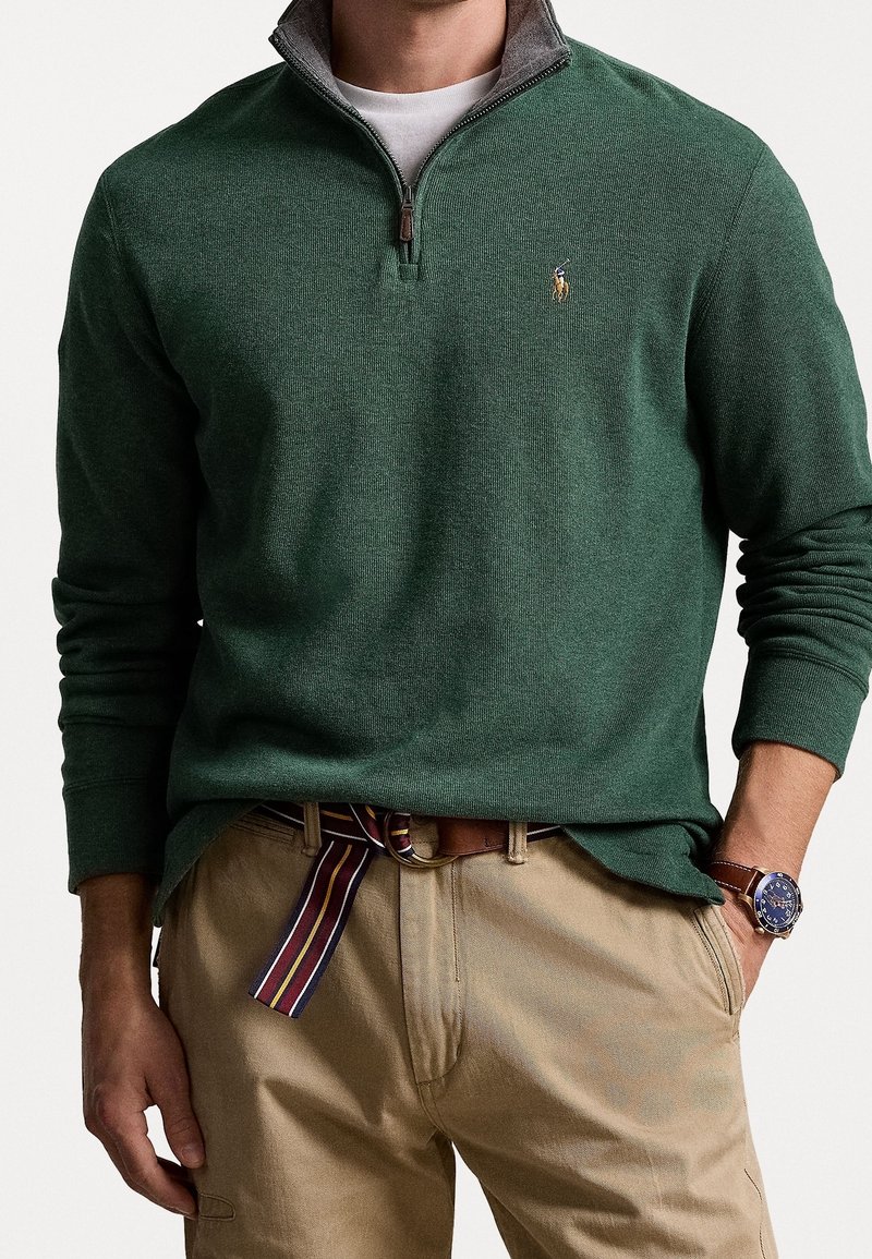 Homme portant un pull vert forêt à fermeture éclair jusqu'au quart, un pantalon beige avec une ceinture en tissu rayé et une montre-bracelet en cuir marron avec un cadran bleu.