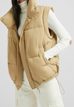 Femme portant un gilet matelassé beige sur un pull à col roulé blanc côtelé et un pantalon blanc cassé, main dans la poche du gilet.