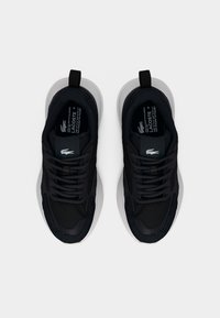 Ζευγάρι μαύρα παπούτσια Lacoste με παχιά λευκή σόλα, μαύρα κορδόνια και λευκά λογότυπα κροκοδείλου στη γλώσσα, εμφανιζόμενα από πάνω σε λευκό φόντο.