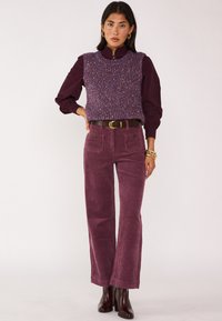 Gebreide paarse vest met multicolore stippen over een donkere bordeauxblouse, gecombineerd met brede corduroy broeken in een lichtere pruimkleur. Bruine riem.