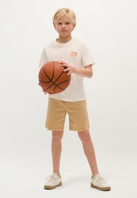 Weißes T-Shirt mit Logo, beiges Shorts und Sneakers, die einen braunen Basketball halten. Minimalistisches Design vor einem schlichten Hintergrund.