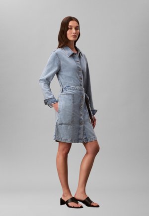 Nő világoskék, hosszú ujjú, gombos farmerruhában, melynek előoldalán zsebek találhatók, fekete, nyitott orrú, magas sarkú szandállal párosítva.