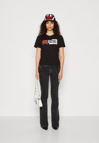 T-shirt en coton noir avec logo « DIESEL » blanc et rouge, associé à un jean noir évasé et un sac à main blanc. Le modèle porte une casquette noir et rouge.
