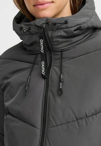 Veste rembourrée grise avec un col haut, cordon de serrage réglable et fermeture éclair noire. Comprend un logo sur la tirette de la fermeture éclair et une texture matelassée.