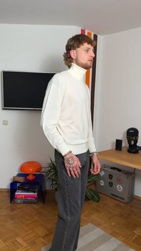 Maglione collo alto color crema con polsini a coste, abbinato a jeans grigio scuro. Una mano mostra tatuaggi dettagliati. Ambiente interno con elementi decorativi.