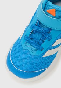 Sinine sportlik toss, millel on võrkmaterjalist pealmine osa, tekstuuriga disain, kontrastsed valged triibud ja erk oranž detail. Varustatud paelte ja polsterdatud kraega.