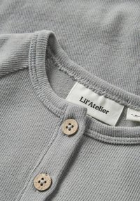 Grå bomullströja med ribbad textur, som har en knäppning framtill med träknappar och en tygetikett som säger "Lil'Atelier."
