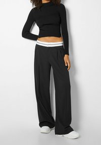 Černý žebrovaný top s dlouhým rukávem a krátkým střihem, doplněný volnými černými kalhotami s bílým páskem s proužky a záhyby. Bílé obuv.