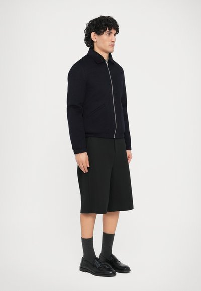 Tengerészkék bomber dzseki cipzárral az elülső részén, gallérral és két oldalzsebbel. Fekete térdig érő rövidnadrág, szürke zokni és fekete mokaszin egészíti ki a szettet.