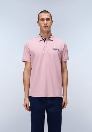 Hombre con polo rosa claro con estampado en el cuello azul marino y pantalones azul marino, de pie frente a un fondo claro y liso.