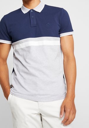 Mężczyzna ubrany w granatowo-szary polo typu color-block z białym kołnierzykiem i białe spodnie, stojący z jedną ręką w kieszeni.