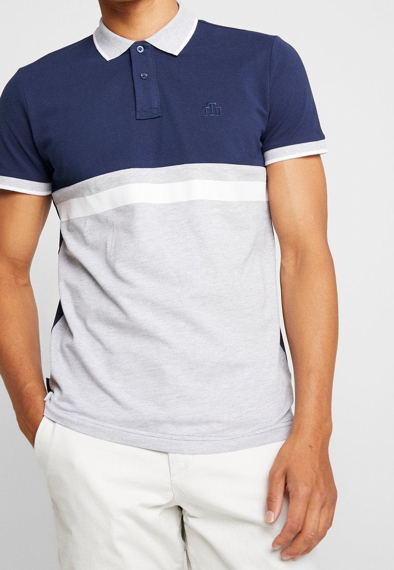 Homme portant un polo bicolore bleu marine et gris clair avec col blanc et pantalon blanc, debout, une main dans la poche.