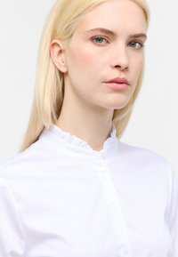 Hvid skjorte med krave, der har en flæsekant, knaplukning og en glat tekstur. Stoffet ser let og sprødt ud.