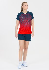 Sporty outfit med en rød til marineblå gradient kortærmet skjorte med et mønstret design, parret med marineblå shorts og hvide sneakers.