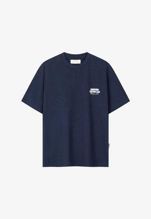 Tums zils īsroku T-krekls ar apaļu kaklu un baltu uzrakstu "ANOTHER COTTON LAB Pilates Lab" kreisajā krūšu pusē.