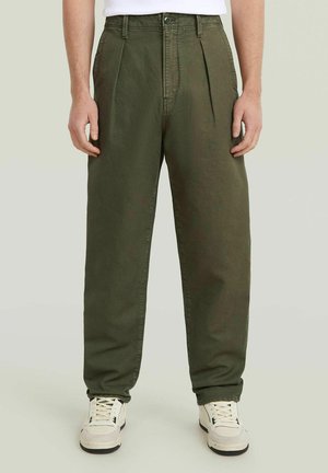Person trägt olivgrüne plissierte Hose und weiße Sneakers und steht vor einem schlichten hellen Hintergrund.