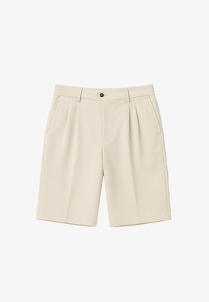 Man draagt beige knielange shorts, witte sokken met zwarte tekst en bruine suède veterschoenen tegen een effen achtergrond.