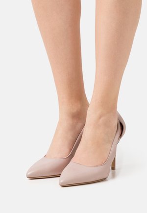 Des pieds portant des escarpins pointus rose pâle se tenant sur une surface blanche.