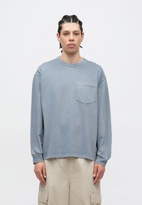 Ανοιχτό μπλε πουκάμισο με μακριά μανίκια, στρογγυλή λαιμόκοψη και τσέπη στο στήθος· κατασκευασμένο από απαλό ύφασμα με άνετη εφαρμογή και ορατές ραφές.