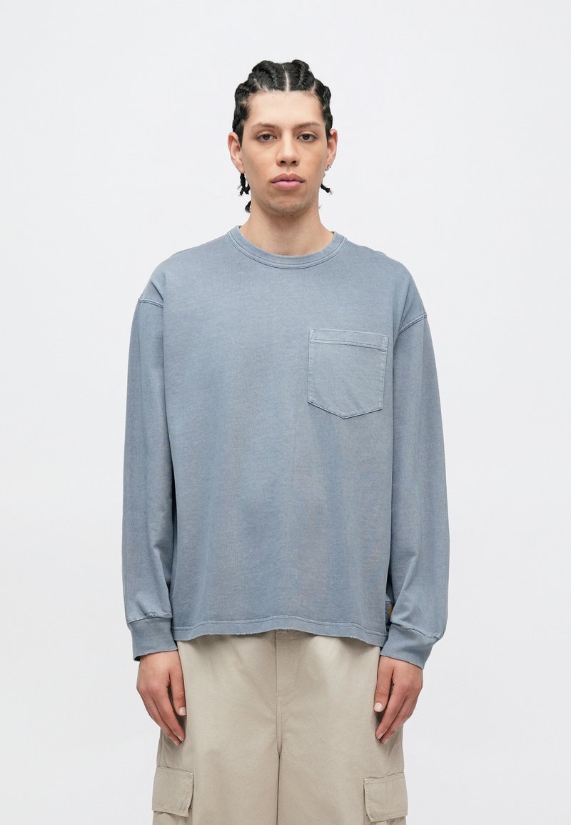 Ljusblå långärmad skjorta med rund hals och bröstficka; tillverkad av mjukt tyg med avslappnad passform och synliga söm-detaljer.