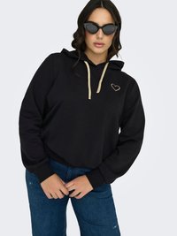 Nainen pukeutunut mustaan huppariin, jossa on kultainen sydäntarra ja kiristysnauhat, tummat aurinkolasit ja siniset farkut, seisoo yksinkertaista vaaleaa taustaa vasten.