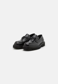 Sorte skinnloafers med en blank overflate, med en tykk svart gummisåle og dekorative spenneakenter foran.