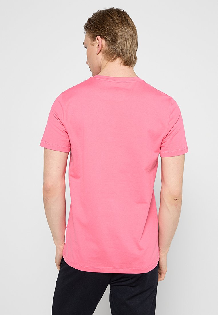 Różowa bawełniana koszulka z krótkim rękawem oraz okrągłym dekoltem, charakteryzująca się prostym wzornictwem i miękką fakturą. Nosi ją osoba widziana z tyłu.