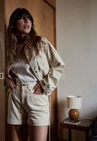 Veste en denim beige et shorts, top à côtes blanches, dans une pièce minimaliste avec des accents en bois et une lampe. Accent sur la texture du tissu et les détails de couleur.