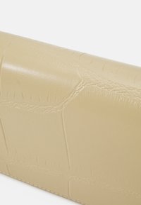 Vaaleanbeige laukku, jossa on krokotiilinnahkajäljitelmä, valkoista taustaa vasten.