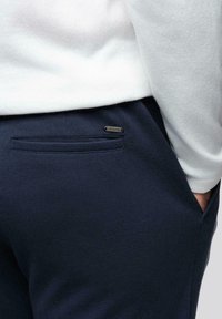 Personne portant un pantalon texturé bleu marine avec une poche arrière passepoilée et une petite étiquette de marque en métal, un haut à manches longues blanc, main dans la poche avant.